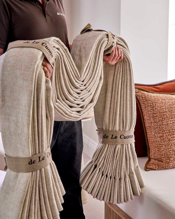 Person holding a large beige blanket with 'de Le Cuona' branding in a minimalistic setting.