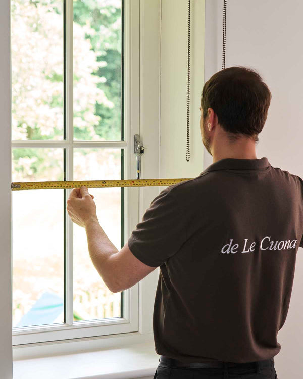 Person measuring a window with a tape measure, wearing a 'de Le Cuona' branded shirt.
