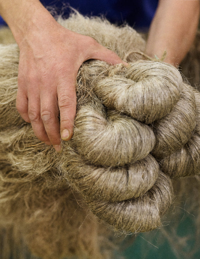 A close-up of a person's hands holding a thick, coiled bundle of natural fiber yarn or wool in a warm beige and gray tone.