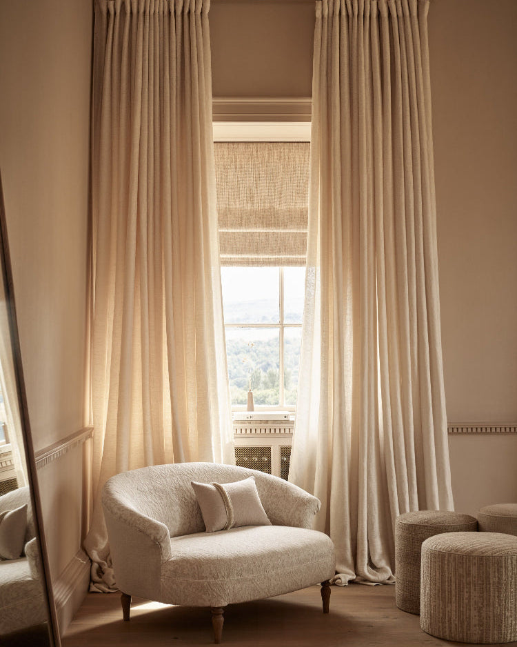 A pair of floor-length beige curtains hanging on a dark rod over a window in a sunlit room, with a cream upholstered armchair and two woven ottomans in front.
