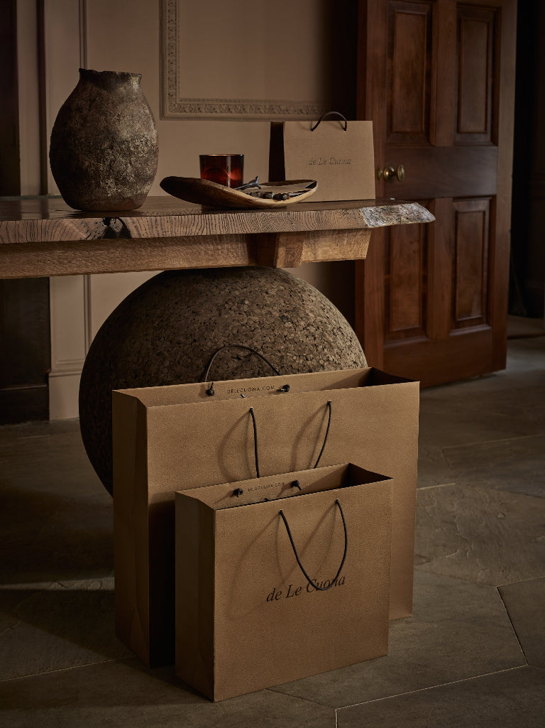 Two brown paper shopping bags with black handles and the printed brand name 'de La Couture' are displayed on a stone floor in a rustic, warm-toned room. A wooden table with a textured vase and other decorative items sits in the background.