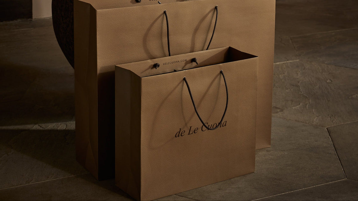 Two brown paper shopping bags with black handles and the printed brand name 'de La Couture' are displayed on a stone floor in a rustic, warm-toned room. A wooden table with a textured vase and other decorative items sits in the background.