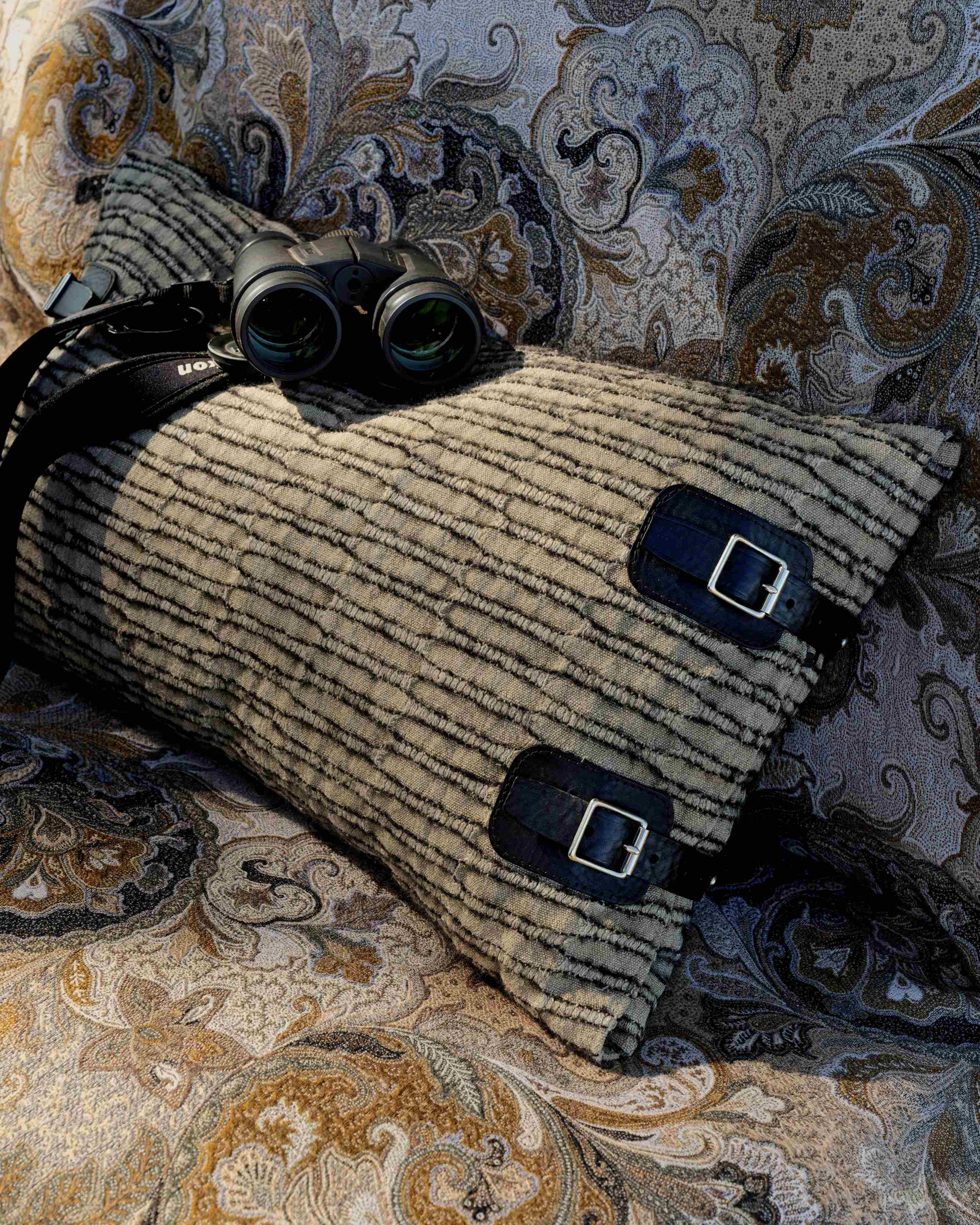 A striped beige and black travel pillow with two black buckle straps, styled on a patterned paisley sofa with a pair of black binoculars resting on top.