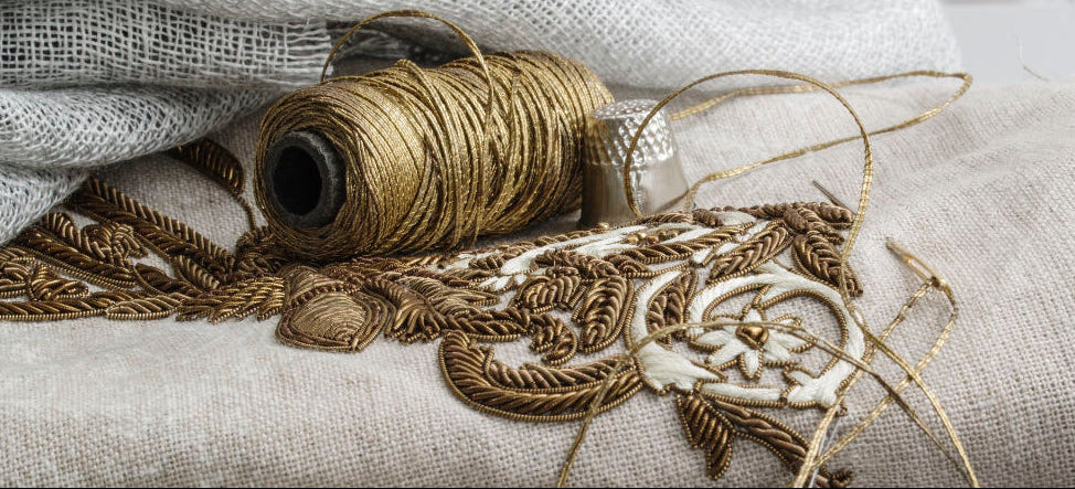 A styled arrangement of folded and draped fabrics in soft neutral tones, including a cream knit textile, light beige linen, a gray woven piece, and a patterned fabric with blue-gray detailing, accented by a spool of tan thread and metallic embroidery.
