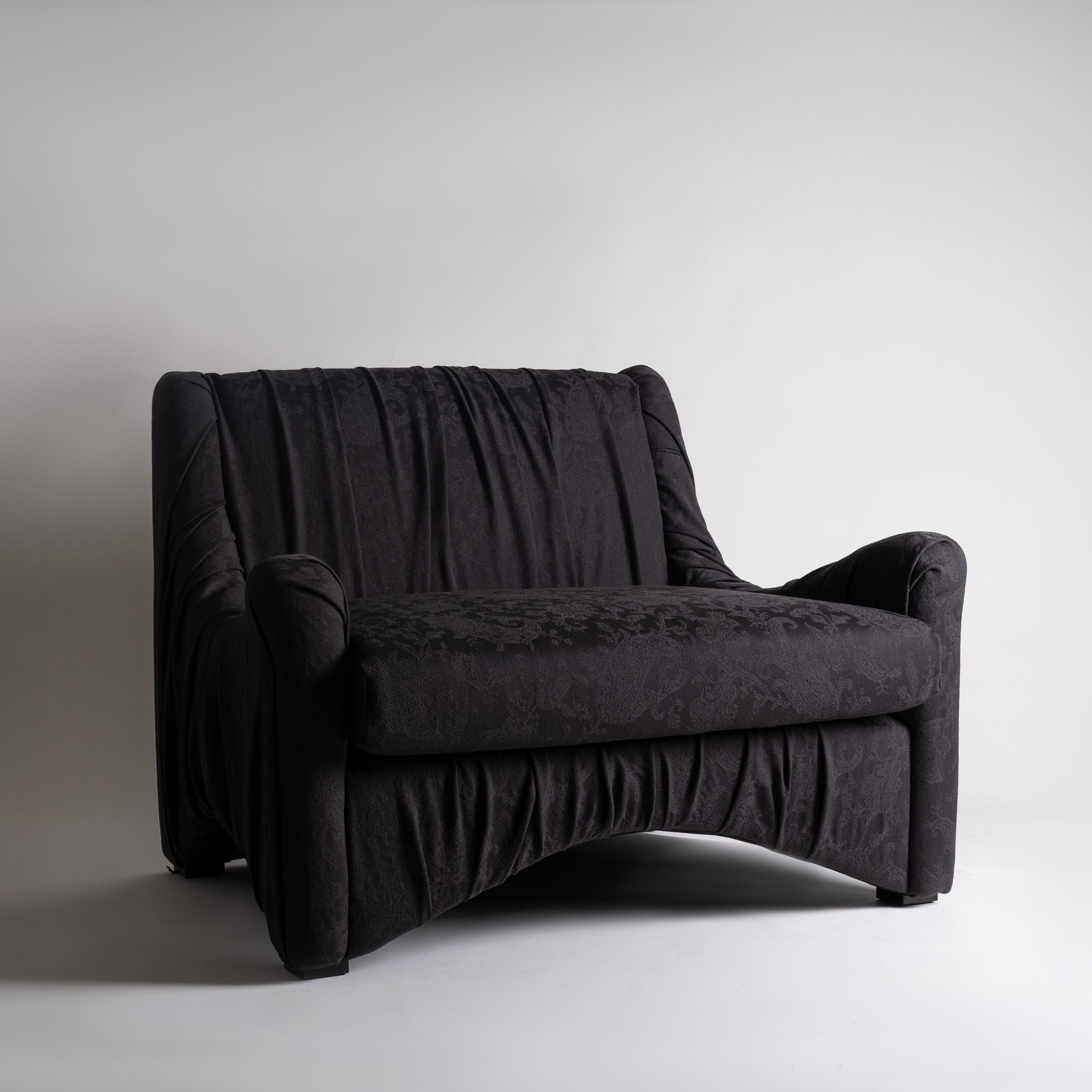 A black upholstered armchair with a heavily padded, draped backrest and curved skirted base, photographed against a plain light gray studio background.