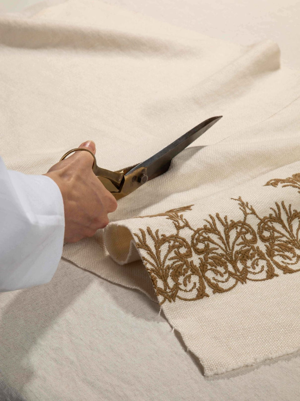 A close-up of a hand holding large scissors and cutting a rolled piece of off-white fabric with ornate gold embroidery along the edge.