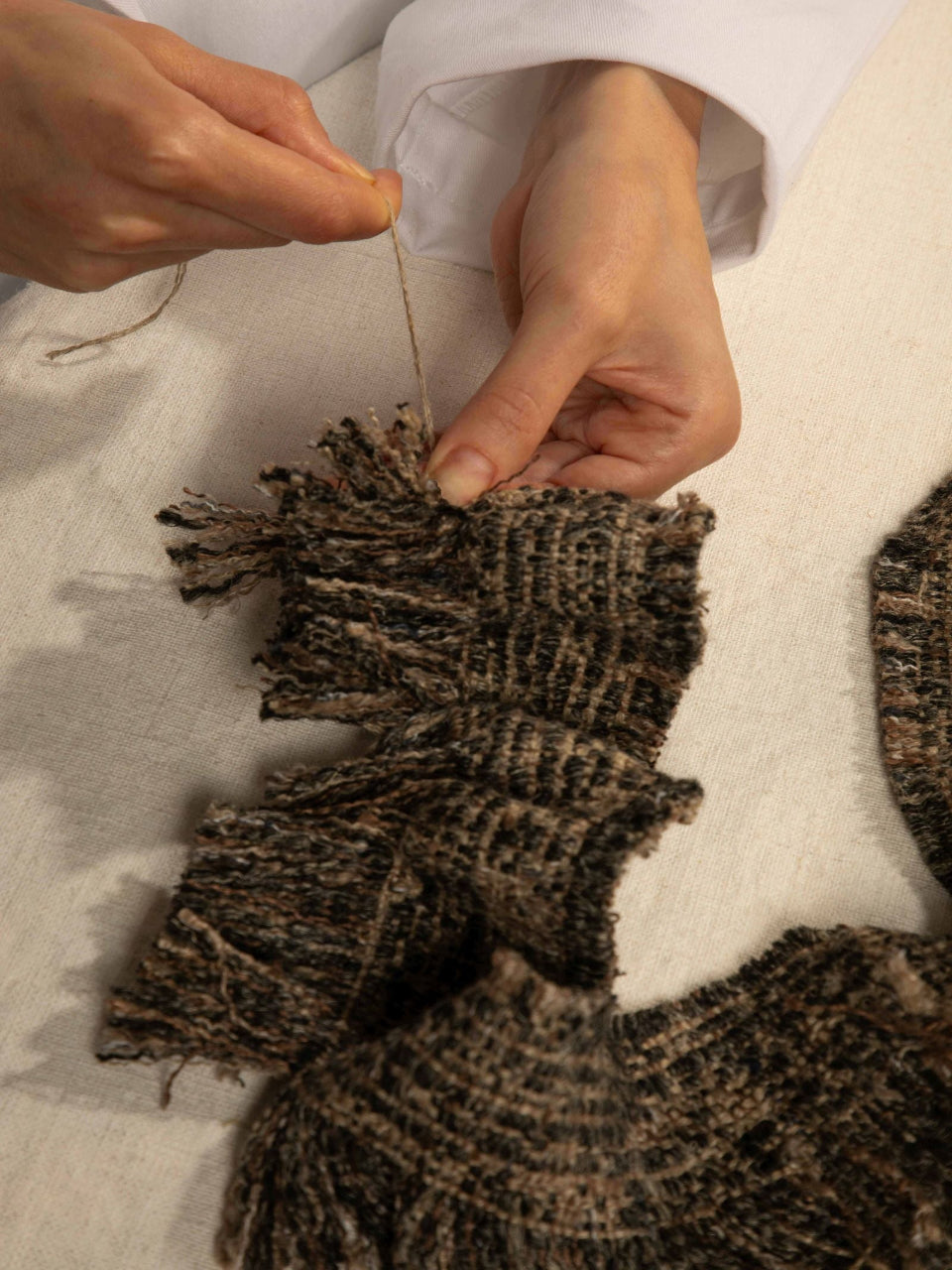 Close-up of a person's hands sewing or stitching a dark brown and black textured fringed textile piece on a light fabric surface.