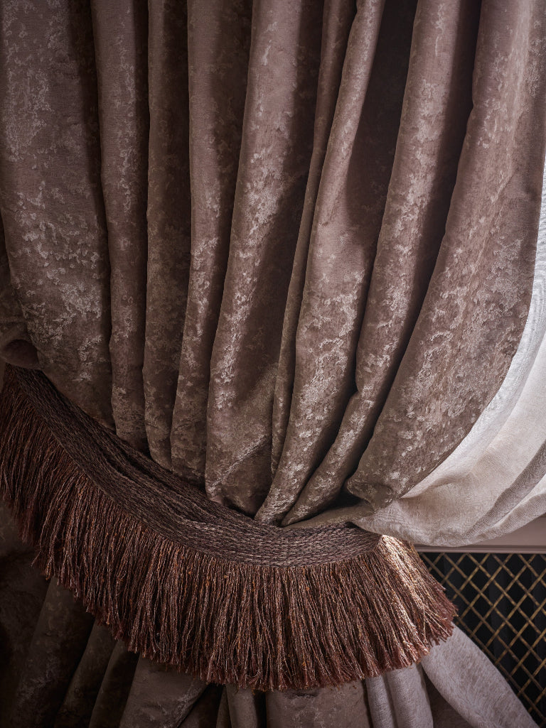 Close-up of a draped curtain in a textured taupe-brown fabric with a matching fringed tieback, next to a sheer off-white inner curtain.