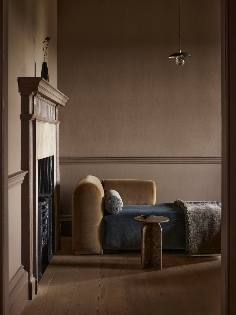 A styled interior scene showing a beige upholstered daybed or chaise with a blue cushion, a round wooden side table, a fireplace with a dark mantel, and warm wood flooring in a dimly lit room.