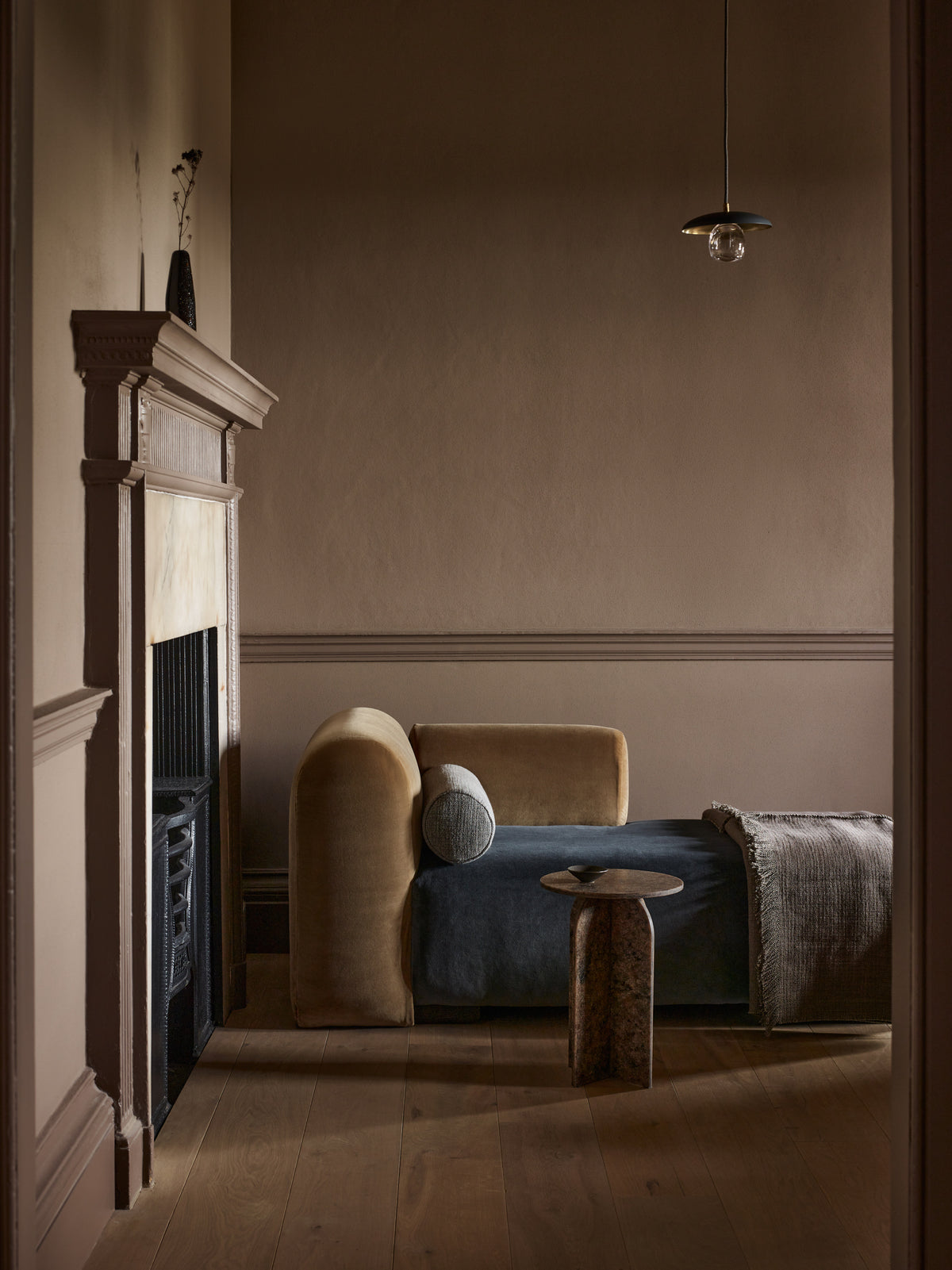 A styled interior scene showing a beige upholstered daybed or chaise with a blue cushion, a round wooden side table, a fireplace with a dark mantel, and warm wood flooring in a dimly lit room.