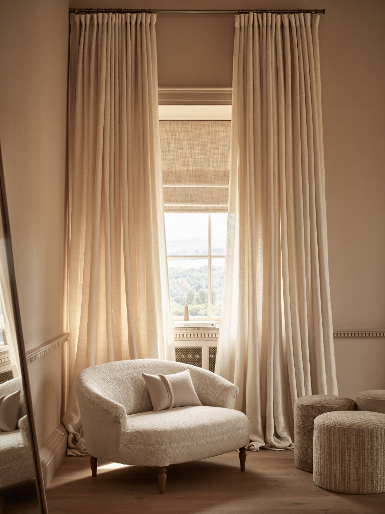 A pair of floor-length beige curtains hanging on a dark rod over a window in a sunlit room, with a cream upholstered armchair and two woven ottomans in front.