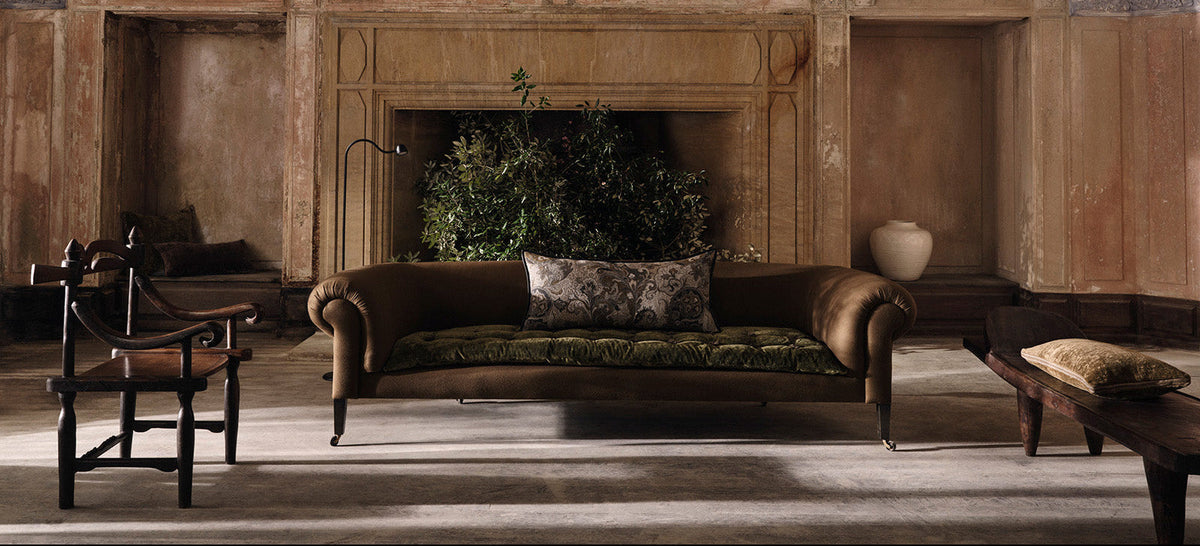 A vintage brown leather sofa with rolled arms and a green patterned cushion, set in a rustic, old-style room with a fireplace and wooden paneling.