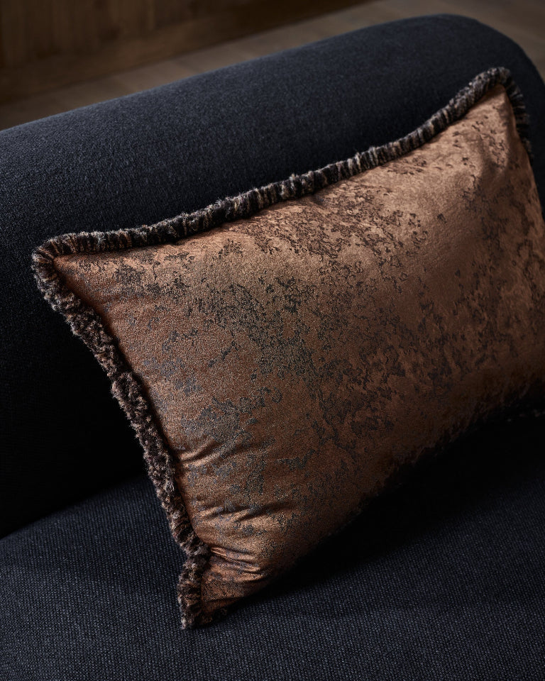A rectangular decorative throw pillow in a distressed bronze-brown fabric with a fringed edge, resting on a dark charcoal-colored sofa.