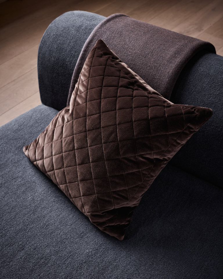 A dark brown quilted velvet throw pillow resting on a dark gray sofa with a matching brown throw blanket draped over the arm, set against a wooden floor background.