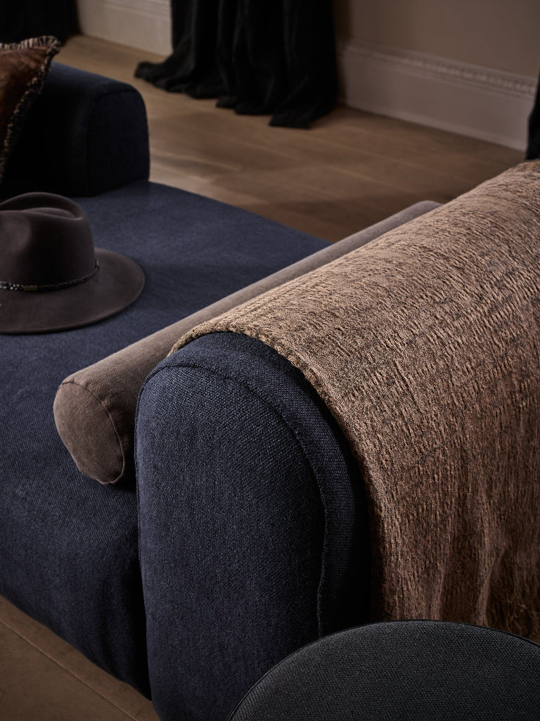 A close-up of a dark navy upholstered chair or sofa with a textured brown throw blanket draped over the arm, plus a brown hat resting on the seat, in an indoor room setting.