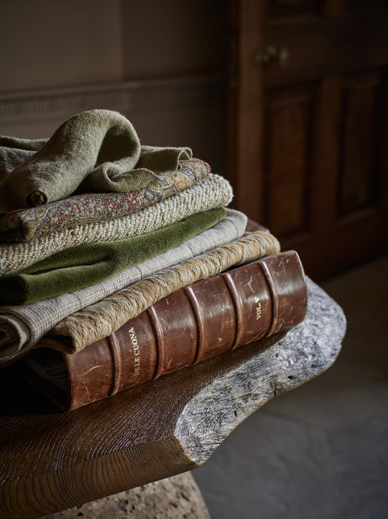 A stack of folded textiles and a vintage leather-bound book resting on a carved wooden table in a dimly lit interior.