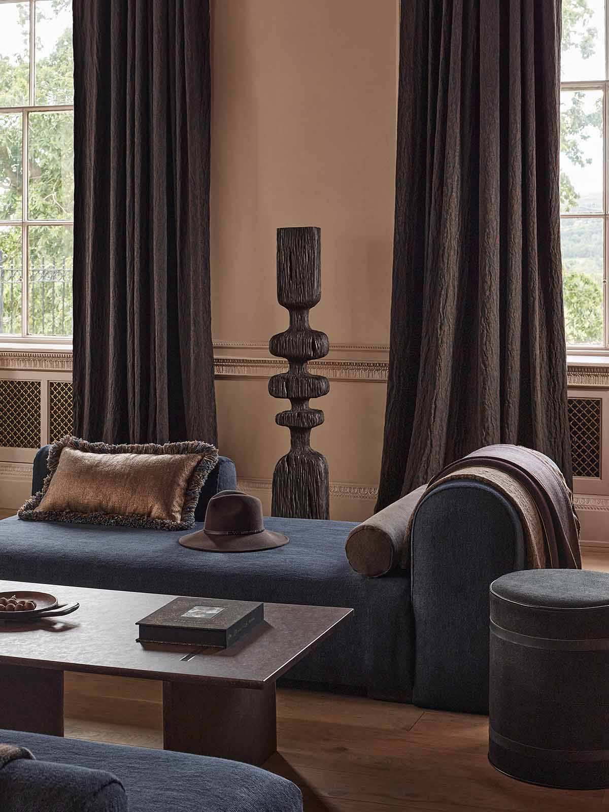 A styled interior room with dark charcoal gray curtains, a blue-gray chaise lounge with cushions, a matching coffee table, and a round ottoman on a wood floor.