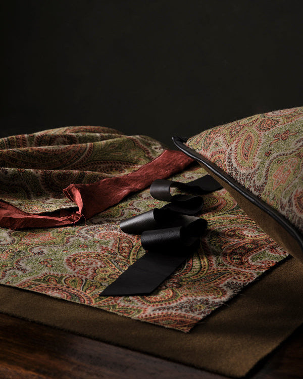 A close-up of a paisley-patterned fabric or scarf in muted green, beige, and rust tones, styled with a black ribbon bow and a dark brown satin ribbon on a dark background.