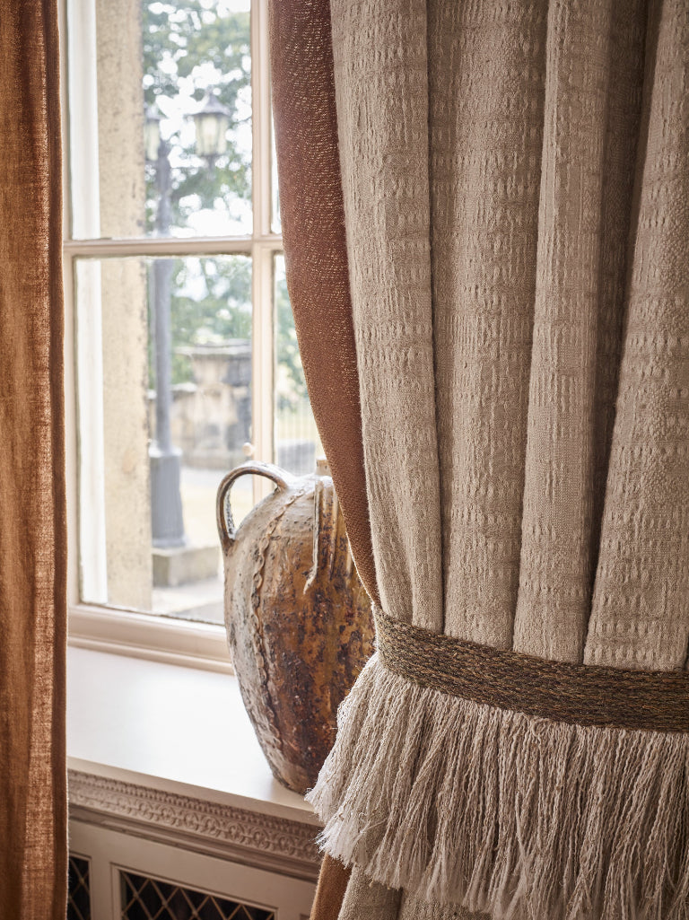 Textured beige curtains with a fringed tieback gathered by a decorative vase on a windowsill.