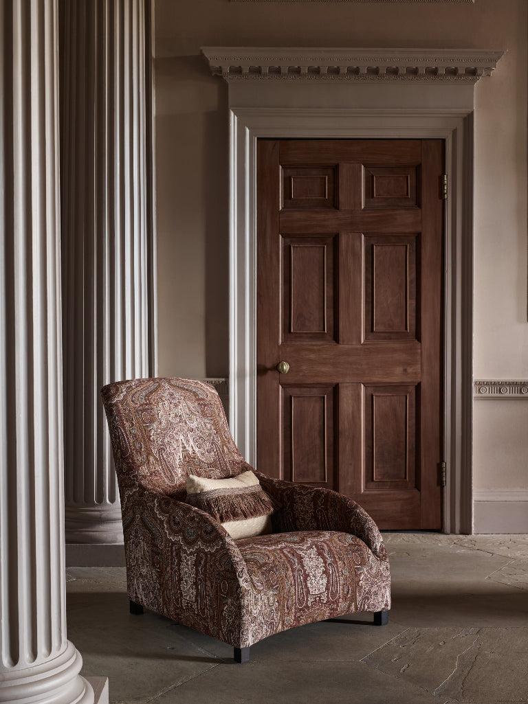 An upholstered armchair with a brown and beige paisley pattern sits in a hallway beside a white fluted column, with a dark wood paneled door in the background.
