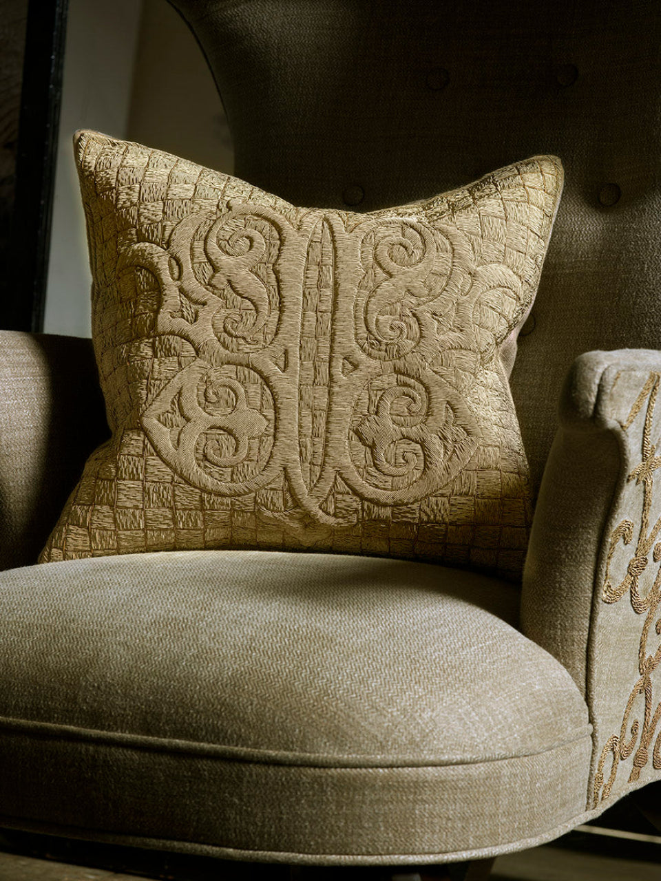 A square beige throw pillow with an ornate embossed scroll pattern, resting on the back of a matching upholstered armchair.