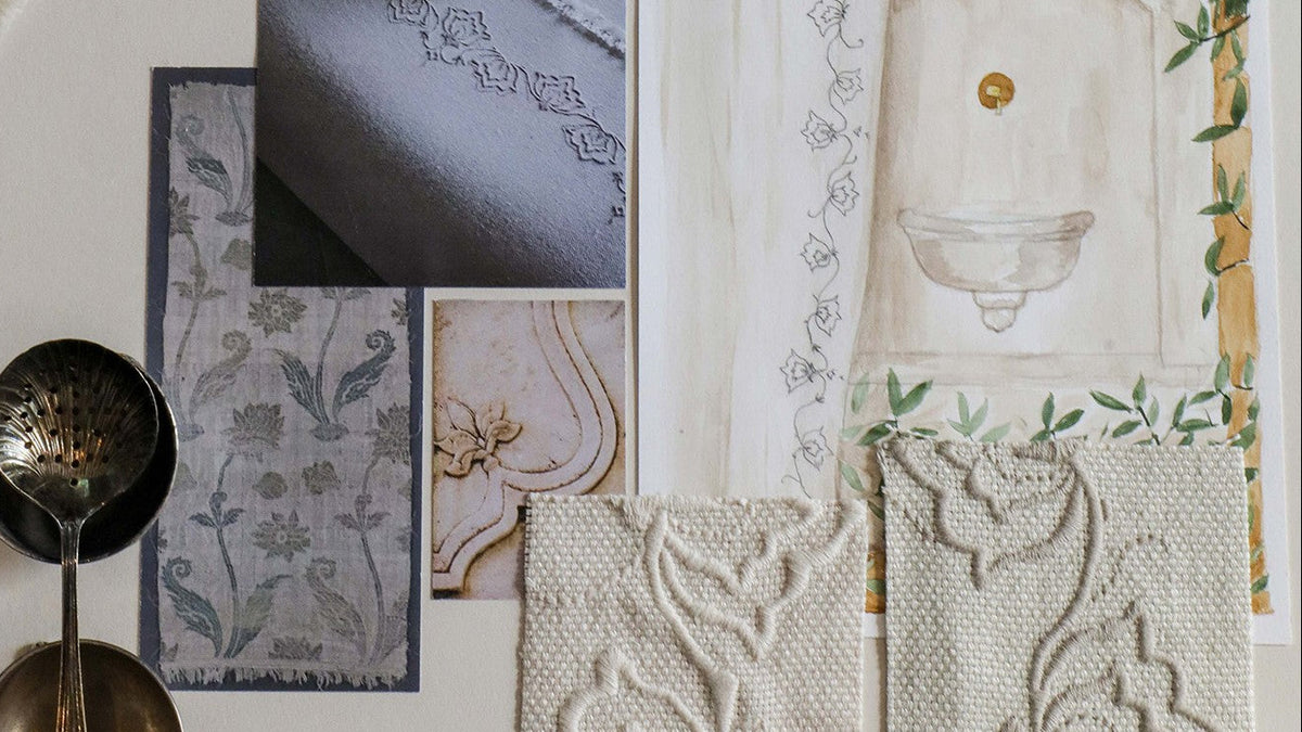 A styled flat-lay of home textiles and decorative accessories on a light neutral surface, including embroidered cream fabric swatches, a floral textile panel, a pillow close-up, a wall-art print with a bowl motif, and two metal tea strainers.