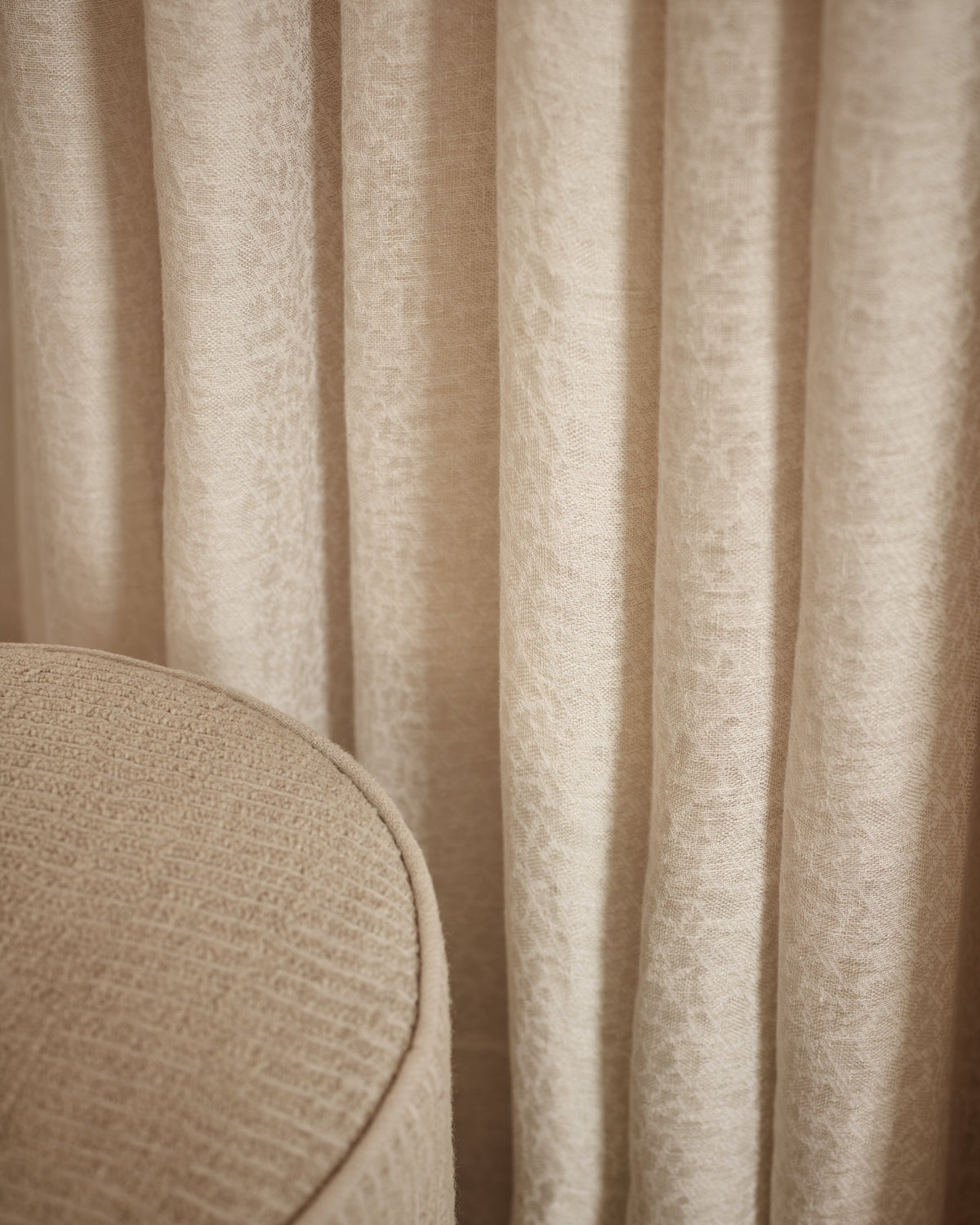 Close-up of textured beige curtains with soft vertical folds and a matching upholstered surface in the lower left corner.