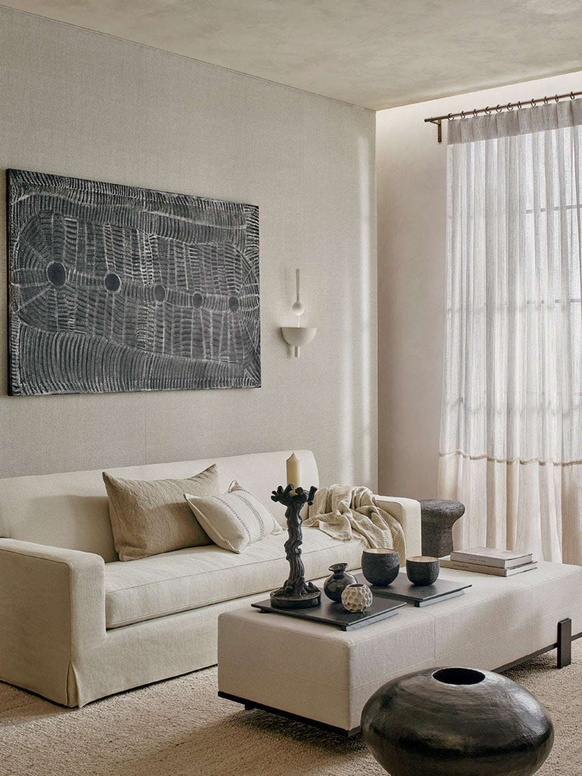 A modern living room featuring a large cream-colored fabric sofa with matching pillows, a low cream coffee table with decorative ceramic vessels, and a large black and gray abstract canvas print on the textured beige wall above.