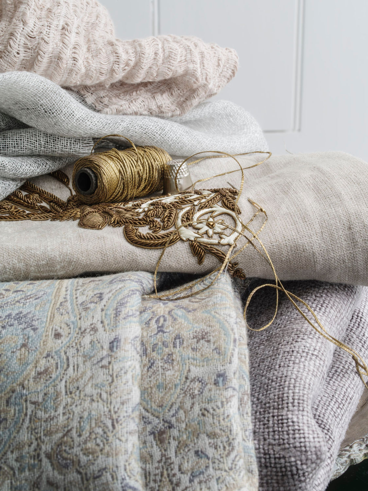A styled arrangement of folded and draped fabrics in soft neutral tones, including a cream knit textile, light beige linen, a gray woven piece, and a patterned fabric with blue-gray detailing, accented by a spool of tan thread and metallic embroidery.