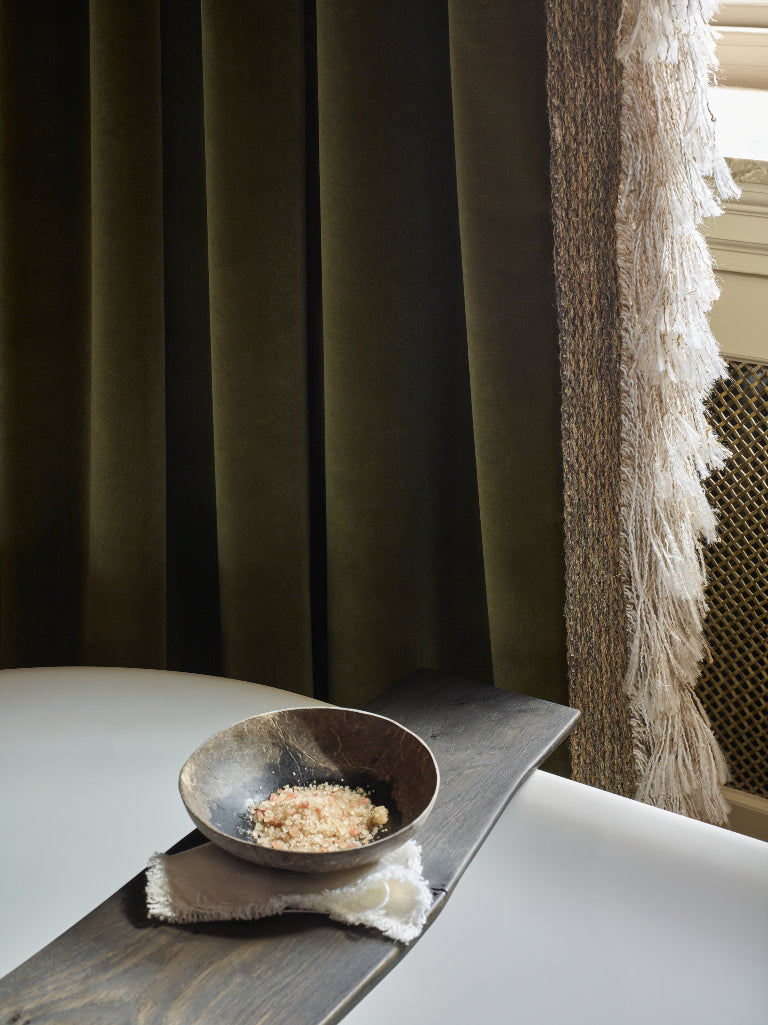 Dark olive green velvet curtains hanging next to a window with a white table in the foreground holding a small rustic bowl.