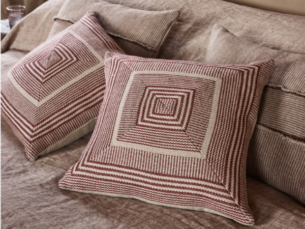 Two square throw pillows with a geometric woven pattern in rust red and cream, styled on a matching textured sofa.