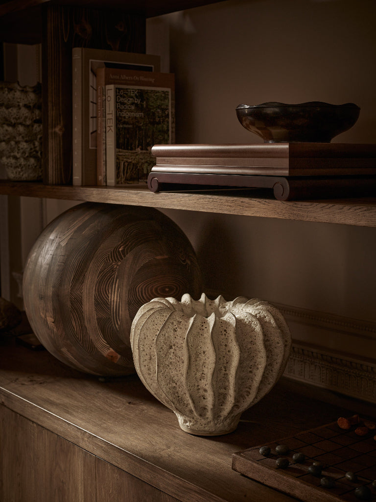 A warmly lit wooden shelf displaying curated home decor. Stacked design books support a dark bowl on the upper shelf, while a large textured wooden vessel and a cream-colored sculptural ceramic vase sit on the lower shelf. The scene features rich wood tones and soft shadows.
