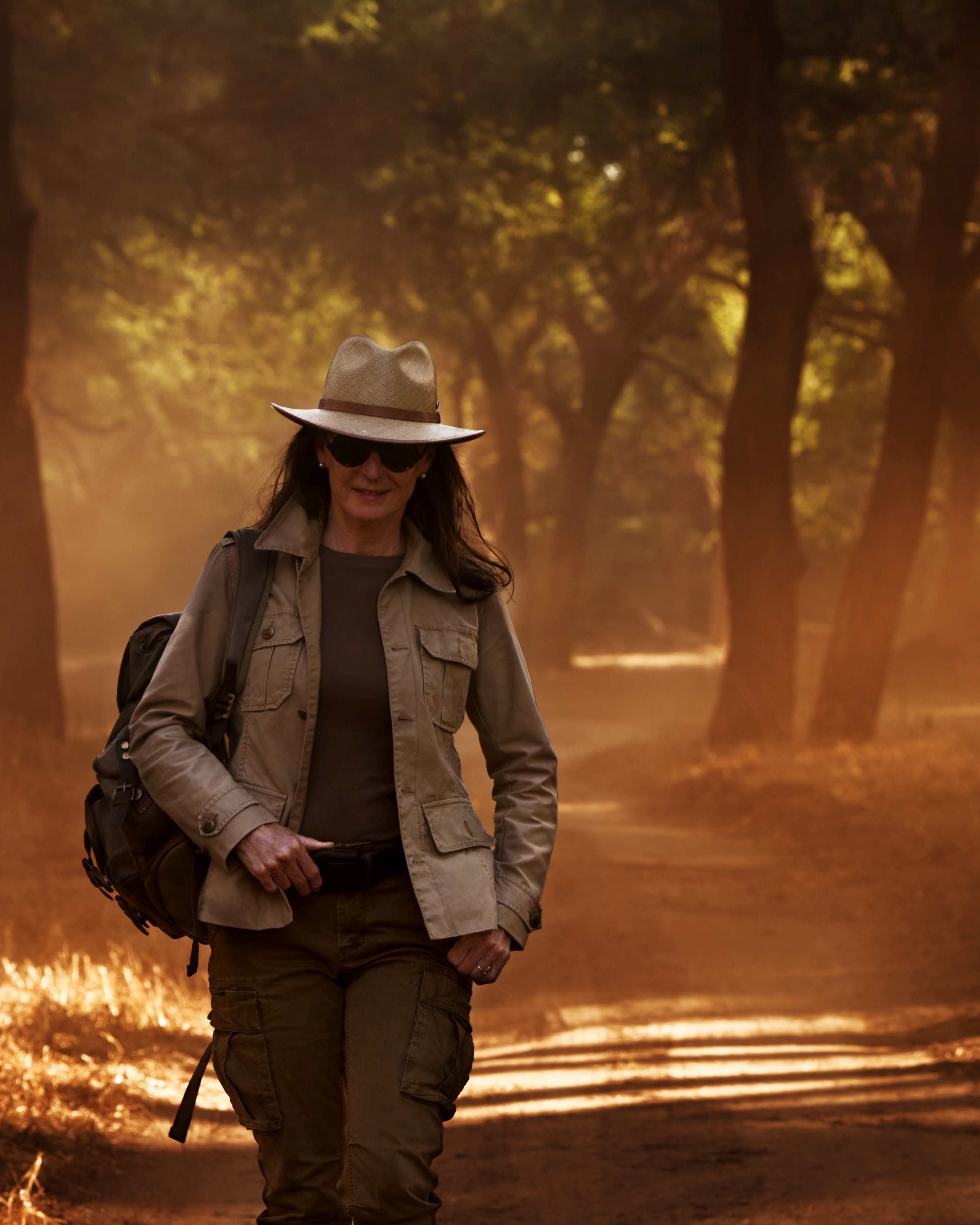 A woman wearing a safari-style hat, olive jacket, and backpack walks along a sunlit forest path at golden hour.