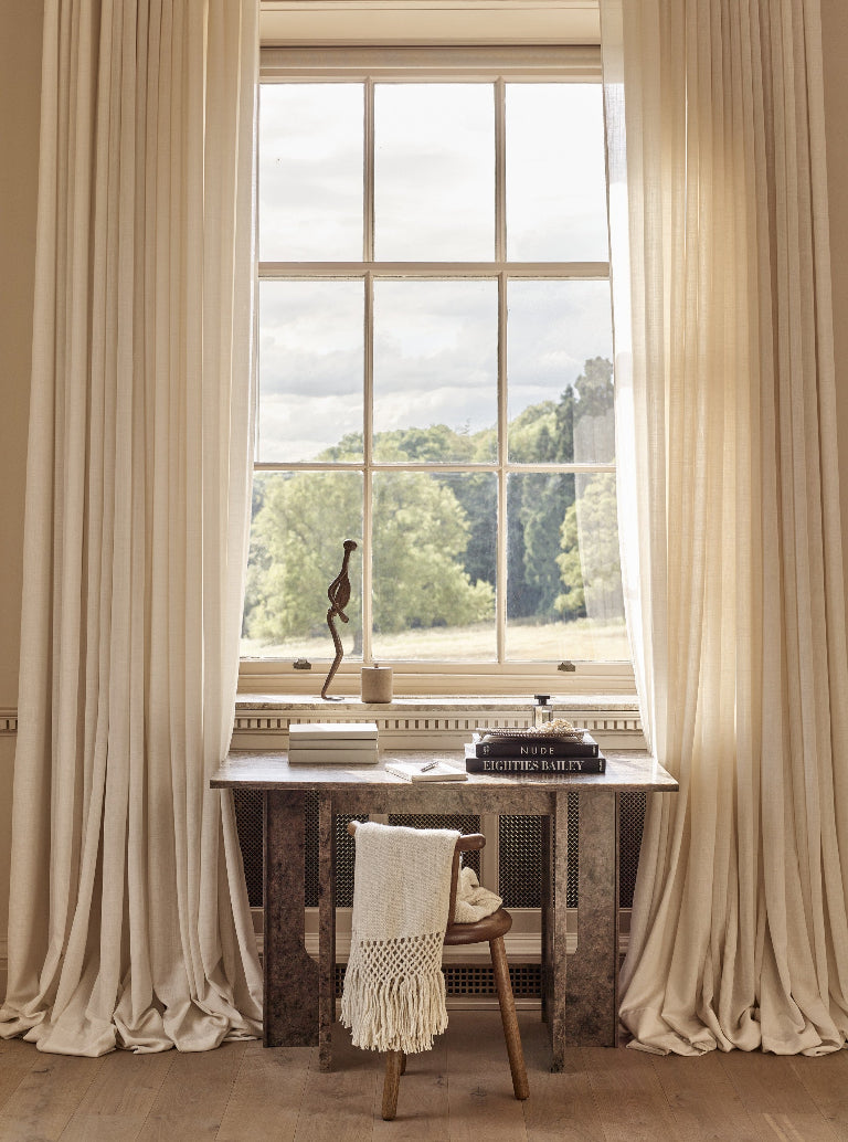A pair of floor-length cream drapes framing a large multi-pane window in a sunlit room, with a small table and chair beneath the window and a view of green trees outside.