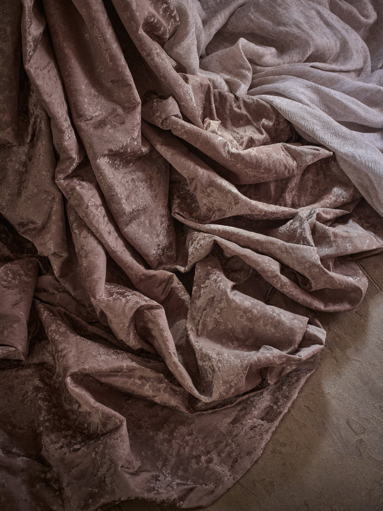 Rumored velvet fabric in a dusty mauve-brown color, draped in soft folds over a neutral concrete floor.