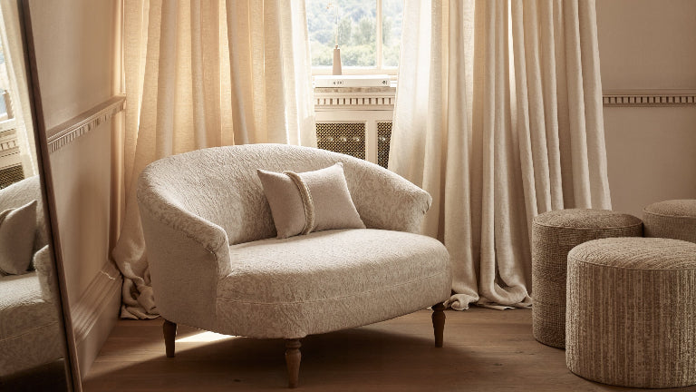 A pair of floor-length beige curtains hanging on a dark rod over a window in a sunlit room, with a cream upholstered armchair and two woven ottomans in front.