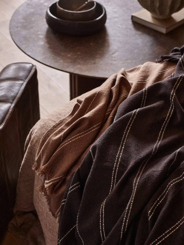 A close-up of draped textiles in a styled interior setting, featuring a dark brown throw blanket with thin light vertical stripes and a tan woven throw draped over a chair beside a round dark table.
