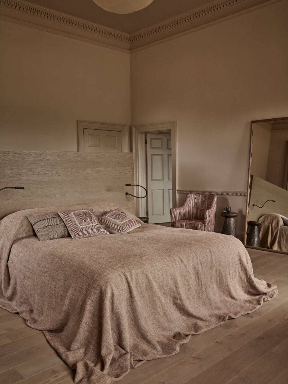 A rustic, minimalist bedroom featuring a large bed dressed in a textured beige linen coverlet and matching pillows. The room has warm beige walls, light wood flooring, a wooden headboard, and a large floor mirror reflecting the bed.