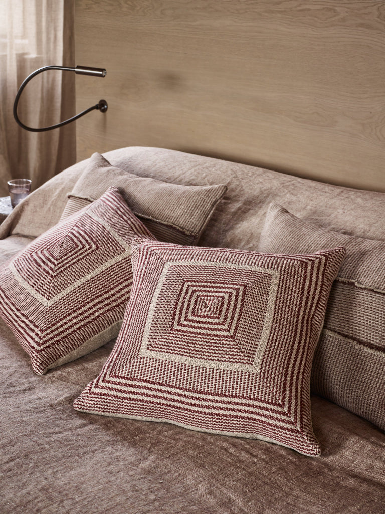 Two decorative throw pillows with geometric striped patterns in rust red and cream, styled on a bed with matching textured bedding and a wooden headboard.