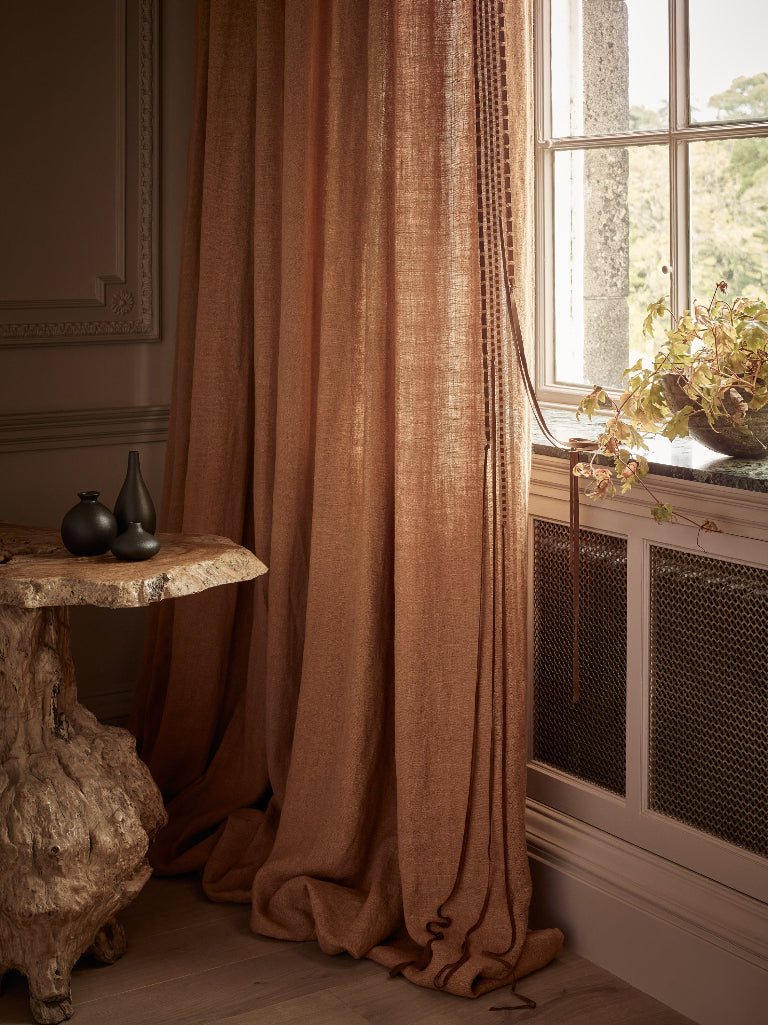 A pair of floor-length rust-colored linen curtains hanging by a window in a styled room with a rustic wooden side table and a potted plant on the sill.