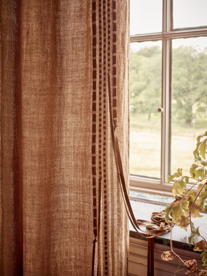 A close-up of a textured brown curtain panel hanging next to a window, with a decorative vertical stripe detail and a plant visible outside.
