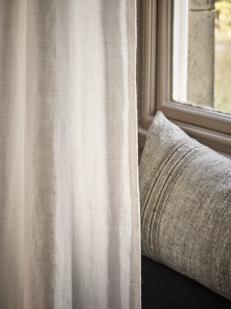 A close-up of a light beige linen curtain hanging next to a window, with a textured gray throw pillow resting on a dark window seat.