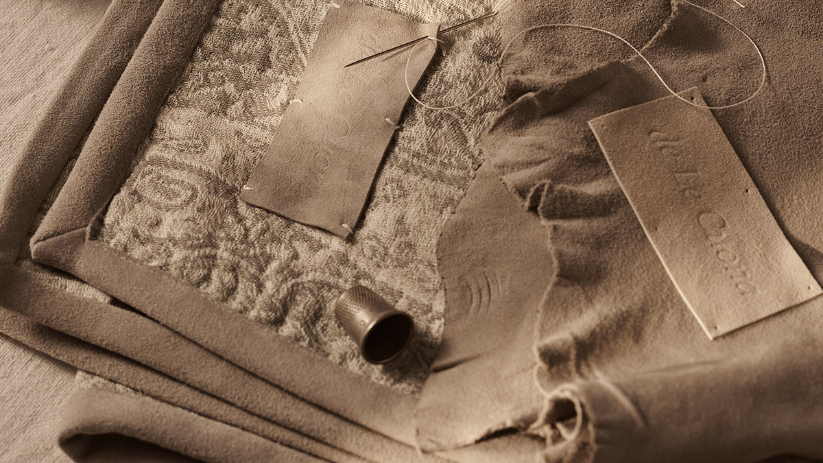 Folded beige and taupe patterned fabric pieces with leather-like labels and ribbons, arranged on a textured neutral surface.
