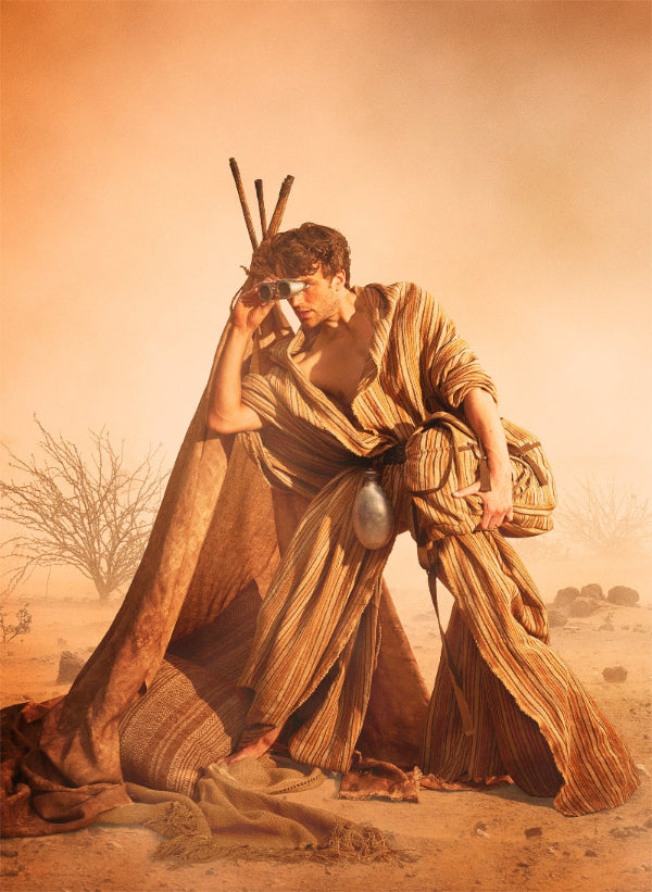 A man in a desert setting wearing a flowing striped brown and beige robe, holding a metal container and standing beside a large draped textile. The scene is bathed in warm orange light with a dusty, atmospheric background.