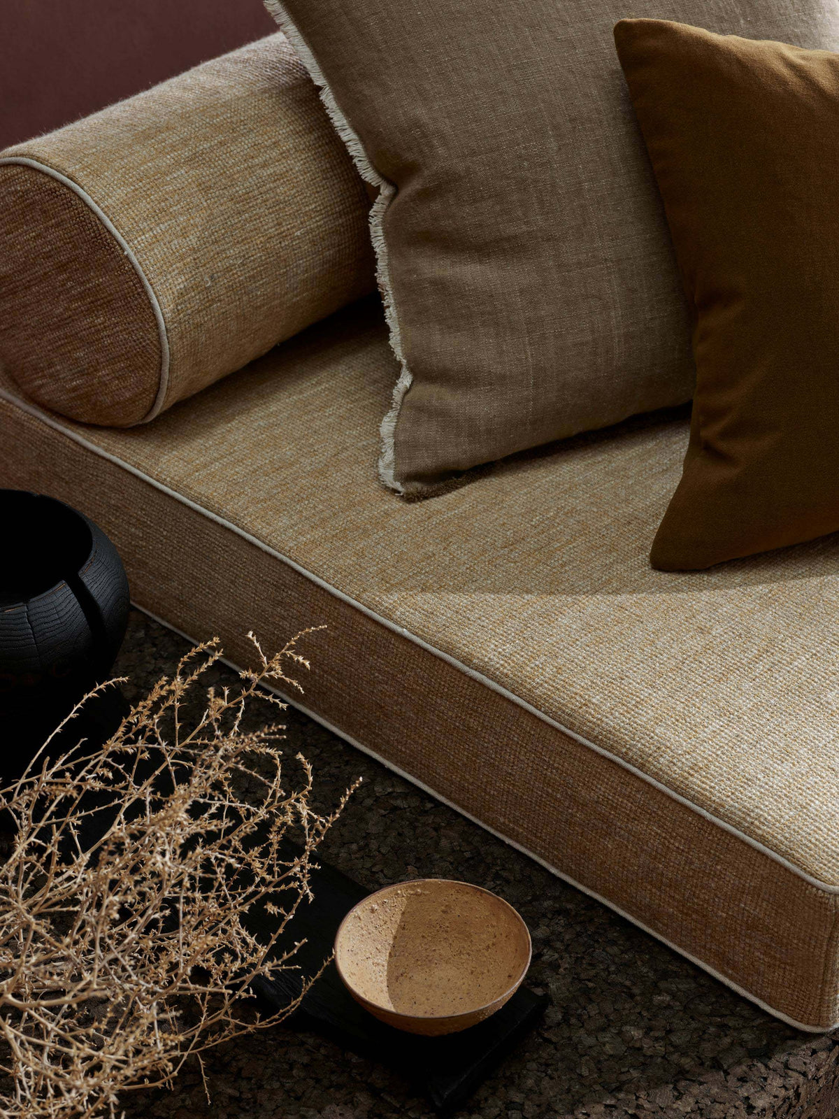 A low-profile upholstered daybed or sofa in warm beige linen fabric with contrasting dark brown piping, styled with matching beige and dark brown pillows, dried branches, and a small bowl on a dark textured floor.