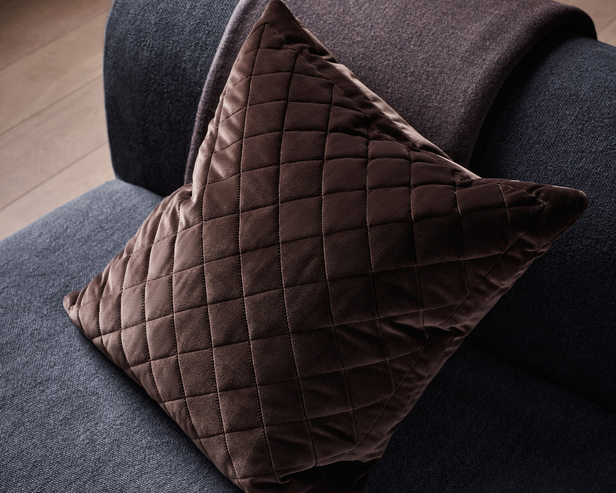 A dark brown quilted velvet throw pillow resting on a dark gray fabric sofa, with a matching dark gray throw blanket draped over the arm.