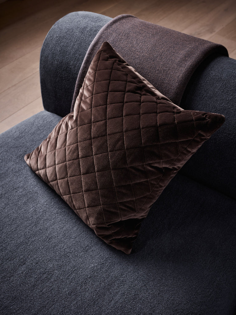 A dark brown quilted velvet throw pillow resting on a dark grey sofa with a matching brown throw blanket draped over the arm, set against a wooden floor background.