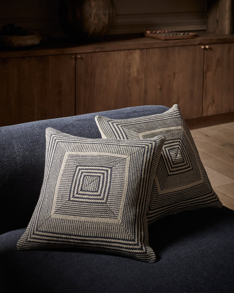 Two square throw pillows with a geometric woven pattern in beige and dark charcoal, styled on a dark blue sofa in a warm, dimly lit living room setting.