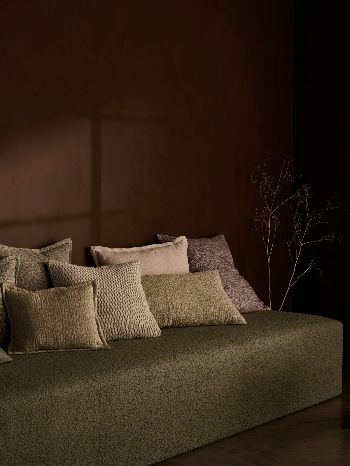 A low upholstered bench or daybed in dark olive green, styled with multiple textured and solid throw pillows in beige, taupe, and mauve tones against a dark brown wall.