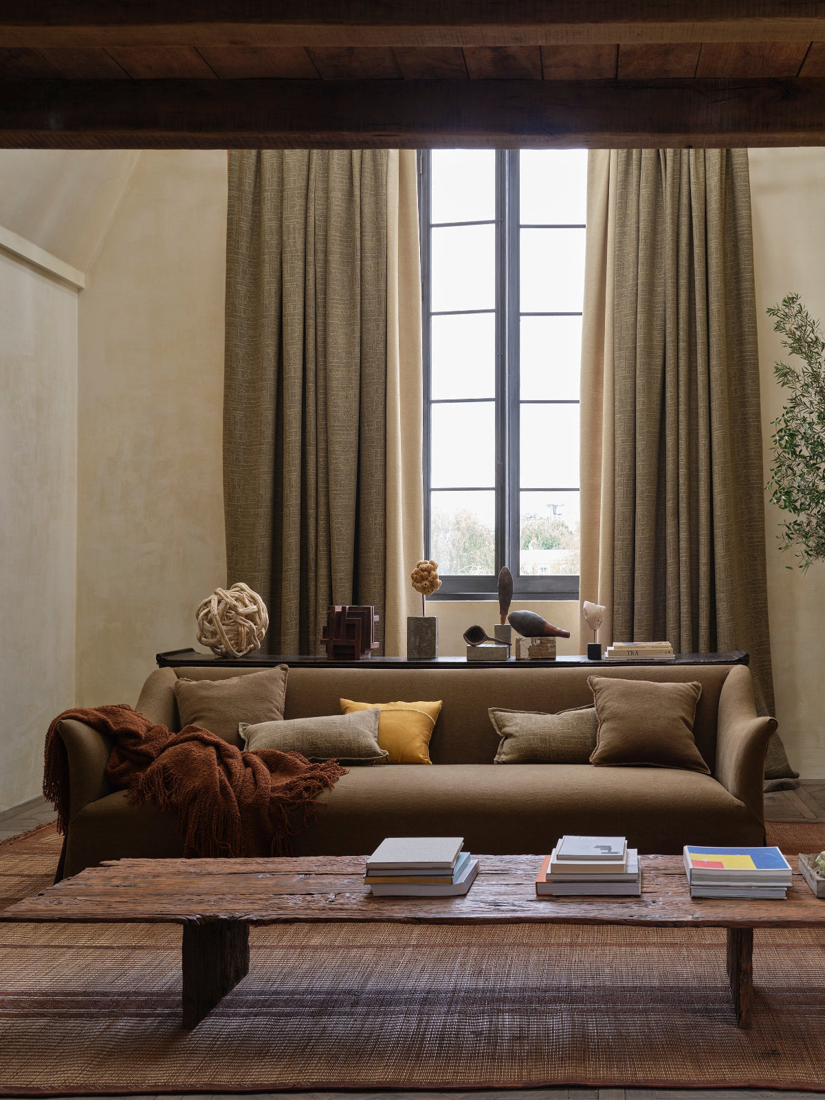 A styled living room interior featuring a brown sofa with neutral and yellow accent pillows, a low wooden coffee table, a large woven rug, beige walls, a tall window with drapes, and a potted olive tree.